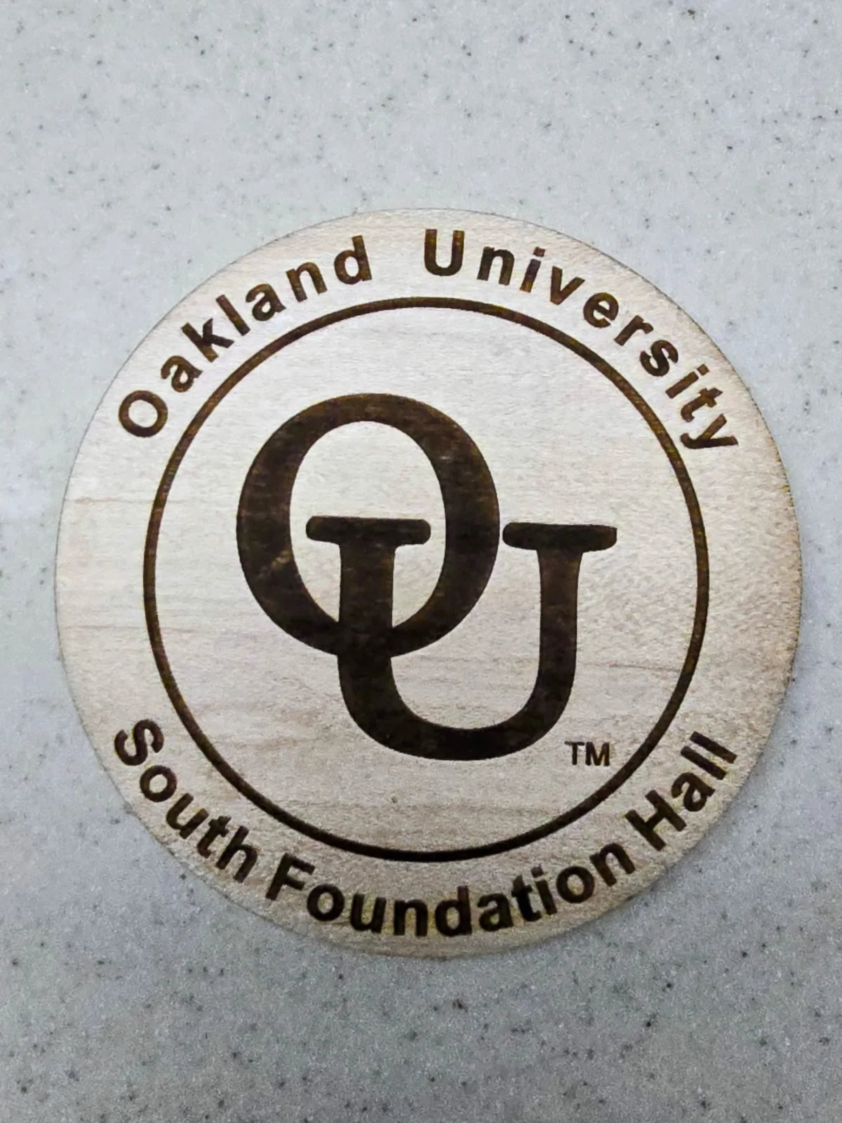 The Oakland University South Foundation Hall coaster in daylight, the engraved wordmark and OU monogram clearly legible in the maple.