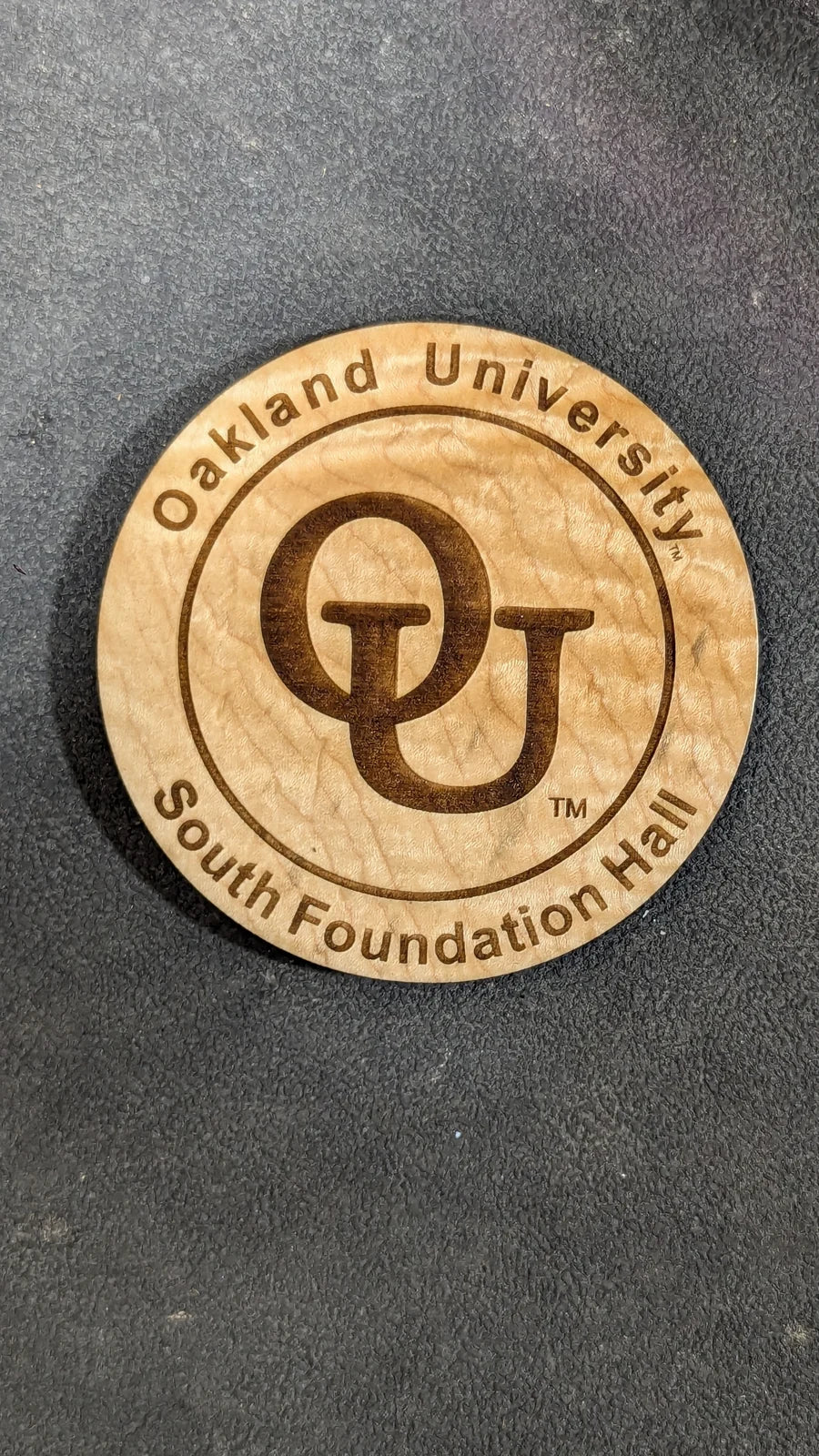 A round engraved coaster reading Oakland University South Foundation Hall.