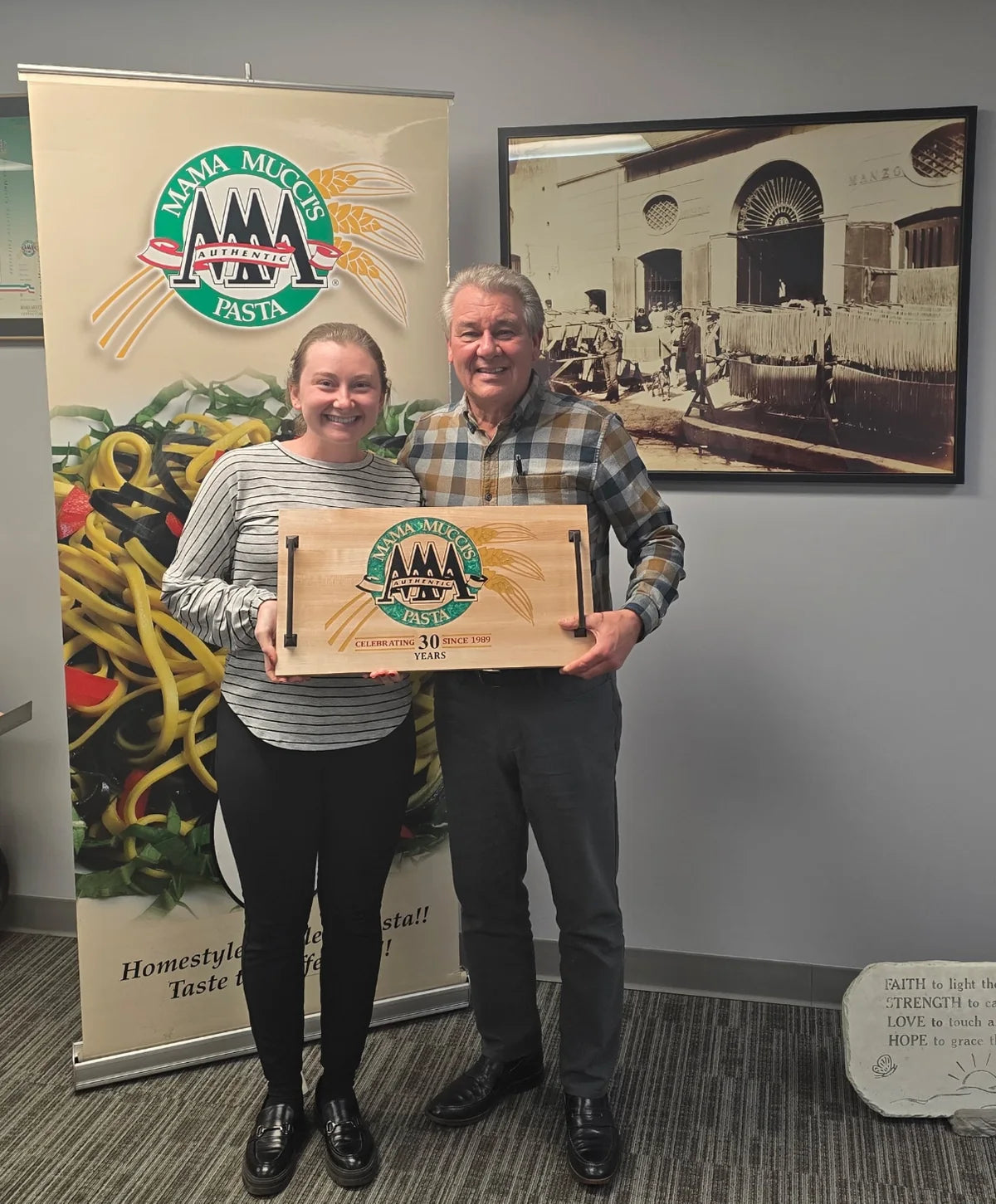 Two people from Mama Mucci's Pasta holding their engraved anniversary board in front of the company banner.